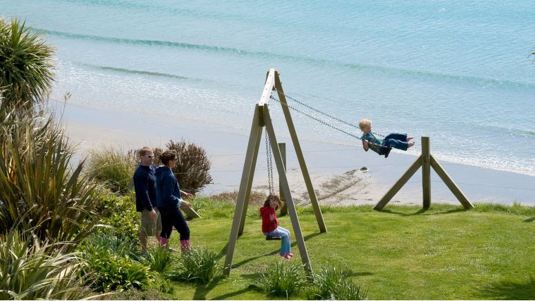 A family plays on the swings at The Nare hotel, with Carne Beach below them.