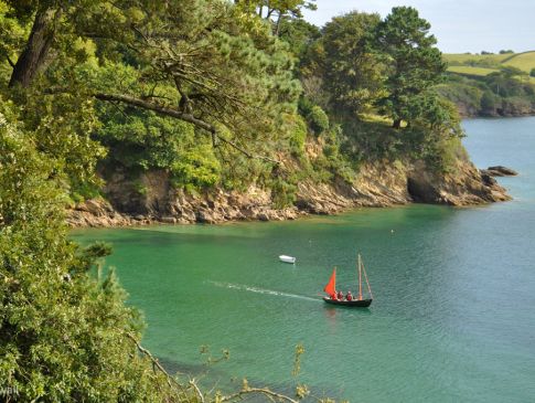 The verdant banks of the Helford River, with a sailing boat gliding over the water.